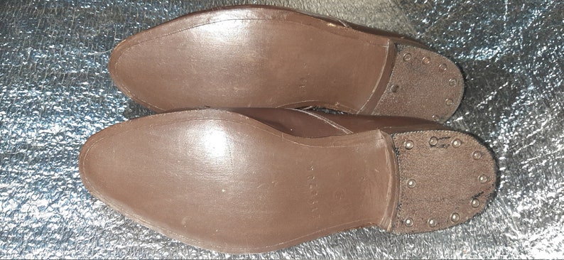 May include: A pair of brown leather shoes, soles facing up. The soles are a solid brown color, with a textured appearance. The heels are reinforced with metal studs.