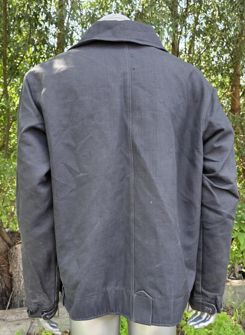 K&ouml;nnte beinhalten: Eine anthrazitfarbene Jacke mit klassischem Kragen und gekn&ouml;pften Manschetten. Die R&uuml;ckansicht zeigt eine taillierte Passform mit einer vertikalen Naht in der Mitte. Die Jacke scheint aus einem strapazierf&auml;higen, m&ouml;glicherweise leinwandartigen Material zu sein.