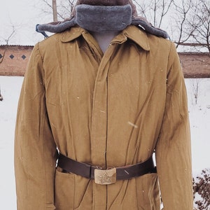 May include: A tan military-style jacket with a brown belt and a star-shaped buckle. The jacket has a matching brown fur hat with ear flaps. The outfit is displayed on a mannequin in a snowy outdoor setting.