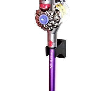 May include: A purple and silver Dyson V8 Animal+ cordless vacuum cleaner is mounted on a black wall mount. The vacuum cleaner is in the upright position with the brush head facing up. The wall mount has the text "Dyson V8 Animal+" printed on it.