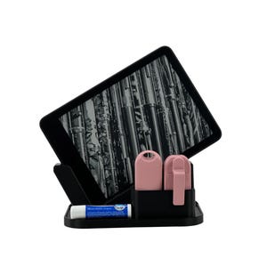 May include: A black desk organiser stand holding a tablet with a black and white flute graphic on the screen. The stand features a small compartment containing two pink rectangular items and a blue and white lip balm tube in front.