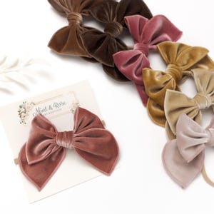 Puede incluir: Un conjunto de seis lazos de pelo de terciopelo en varios tonos de marrón, rosa y beige. Los lazos están atados con un nudo en el centro y tienen una forma ligeramente puntiaguda. Los lazos están dispuestos en una fila sobre un fondo blanco. El texto "Mint & Rose Baby Boutique" es visible en una tarjeta blanca.