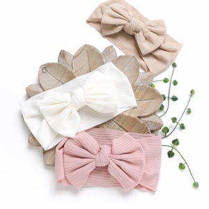 May include: Three ribbed fabric headbands with large bows. The headbands are in beige, white, and pink.