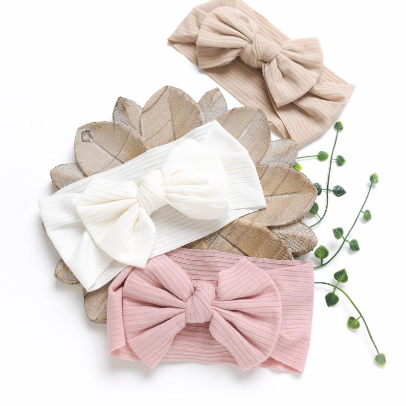 May include: Three ribbed fabric headbands with large bows. The headbands are in beige, white, and pink.