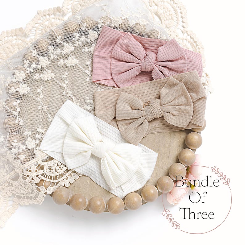 May include: A set of three ribbed headbands with large bows in pink, beige, and white. The headbands are arranged on a wooden tray with a lace background. The text "Bundle Of Three" is visible in the lower right corner of the image.