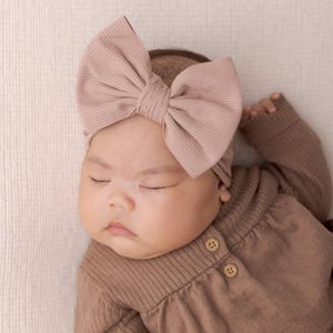 May include: A sleeping baby wearing a brown ribbed knit onesie with two brown buttons and a large pink bow headband.