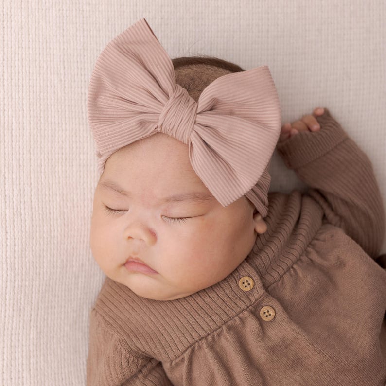 May include: A sleeping baby wearing a brown ribbed knit onesie with two brown buttons and a large pink bow headband.
