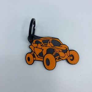 以下が含まれることがあります： オレンジとブラックのキーホルダー。サイドバイサイドのオフロード車のシルエットがデザインされています。