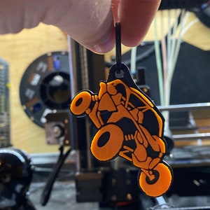 以下が含まれることがあります： オレンジとブラックの3Dプリントされたサイドバイサイドオフロード車のキーホルダー。キーホルダーは黒い紐からぶら下がっています。