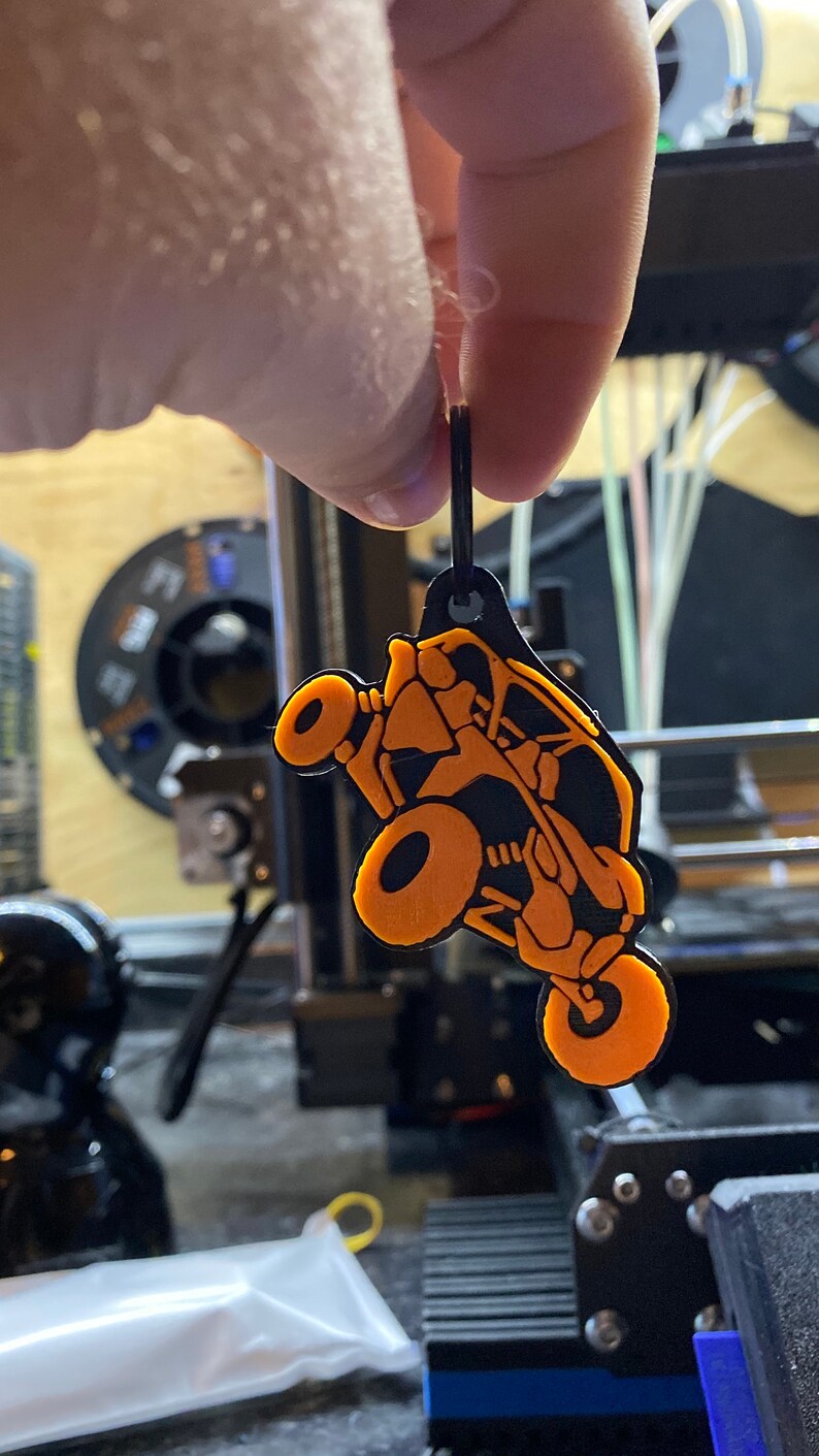 以下が含まれることがあります： オレンジとブラックの3Dプリントされたサイドバイサイドオフロード車のキーホルダー。キーホルダーは黒い紐からぶら下がっています。