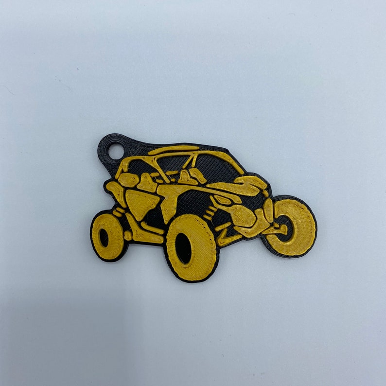 以下が含まれることがあります： 黒と金の3Dプリントされたキーホルダーで、サイドバイサイドのオフロード車の形をしています。