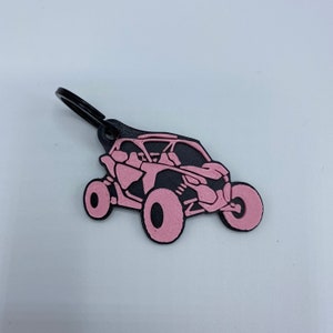 以下が含まれることがあります： ピンクとブラックのキーホルダーで、オフロード車の横顔が描かれています。キーホルダーには黒い金属製のリングが付いています。