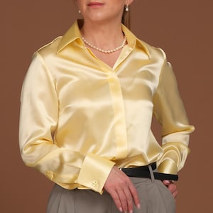 Full-body front view of a woman wearing a luxurious butter yellow silk blouse with French cuffs and a classic collar, styled with elegant wide-leg grey trousers. The smooth, high-quality silk fabric reflects light softly, emphasizing the premium craftsmanship and timeless silhouette of this elegant blouse