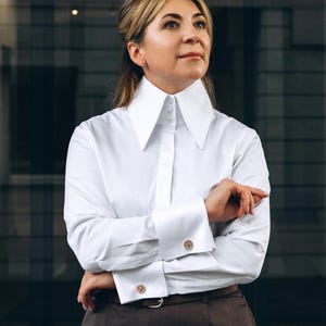 May include: A crisp white long-sleeved button-down shirt featuring an oversized, tall pointed collar and wide double-button cuffs. The shirt is paired with a dark brown skirt secured by a matching belt with a silver-toned buckle.
