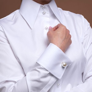 Peut inclure: Chemise blanche à rayures verticales, col classique et boutons nacrés. La chemise présente des poignets français avec de gros boutons de manchette en perles. Les manches sont longues et la chemise est partiellement boutonnée.