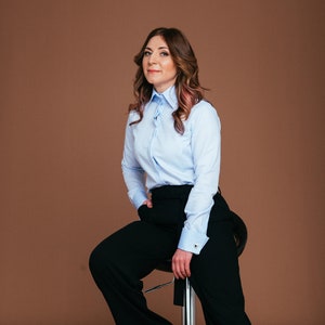 May include: A woman wearing a light blue button-down shirt and black wide-leg pants. The shirt has long sleeves and a classic collar. She is seated on a chrome stool, with one hand in her pocket. Black pointed-toe heels complete the outfit.
