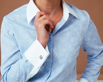May include: A light blue button-down shirt with a white collar and white cuffs. The shirt has a paisley pattern and features silver cufflinks. The shirt is tucked into black pants.