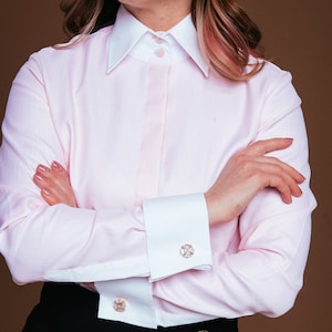 Puede incluir: Una camisa rosa de botones con puños y cuello blancos. La camisa tiene un cierre de botón único en el cuello y dos botones dorados en la parte delantera. La camisa se lleva con una falda negra.