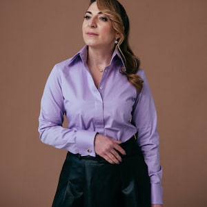 Op de afbeelding: Een vrouw draagt een lavendelkleurig overhemd met knopen en lange mouwen en een zwarte rok. Ze staat voor een bruine achtergrond.