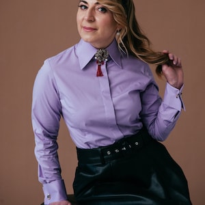 Peut inclure: Une femme portant une chemise boutonnée violette avec une broche décorative et une jupe en cuir noir.