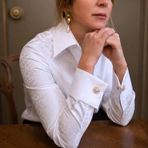 May include: White button-up shirt with a subtle floral pattern. The shirt features French cuffs adorned with gold and crystal cufflinks. A pair of gold earrings with green and white accents are visible.