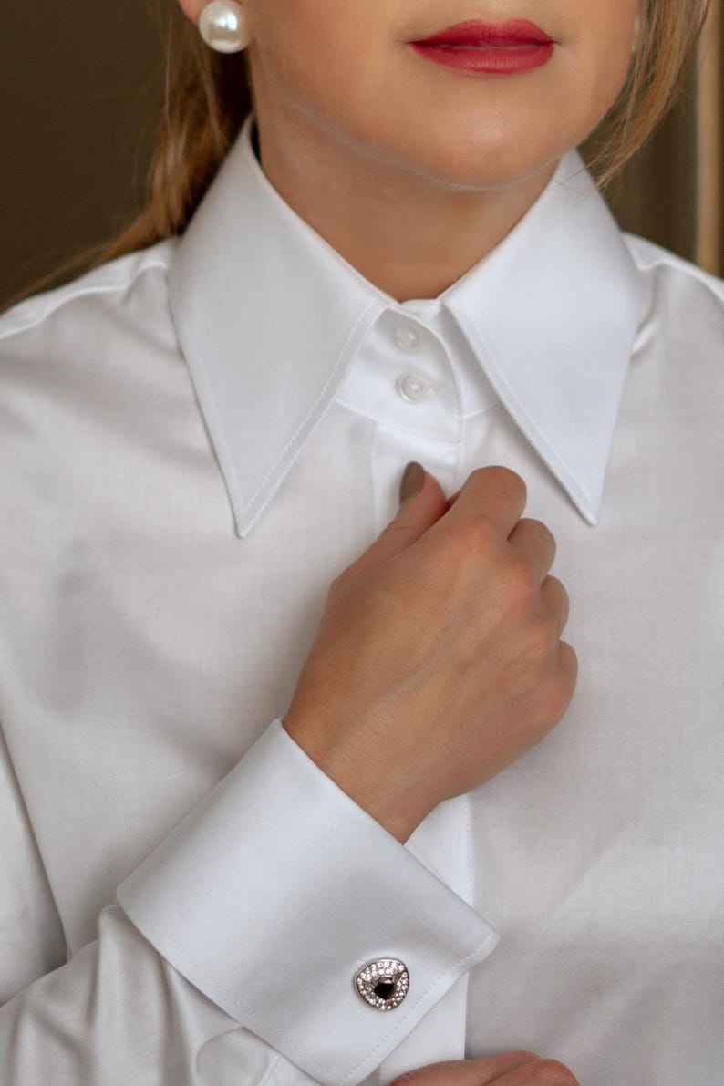 May include: A crisp white dress shirt with a classic collar and French cuffs. The cuff features a silver cufflink with a black center stone and surrounding clear stones. The shirt is buttoned at the collar.