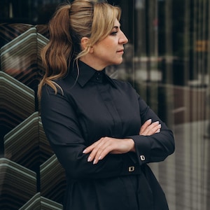 Peut inclure: Une robe chemise noire à manches longues dotée d'un col structuré, d'une patte de boutonnage sur le devant et de poignets ornés de petits détails métalliques carrés. Le vêtement est ajusté à la taille avec un lien à nouer sur le devant.