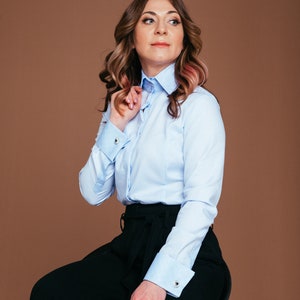 May include: A light blue button-down shirt with a contrasting white collar and cuffs. The shirt is paired with black wide-leg pants. The cuffs feature silver cufflinks. The model is seated on a stool against a brown backdrop.