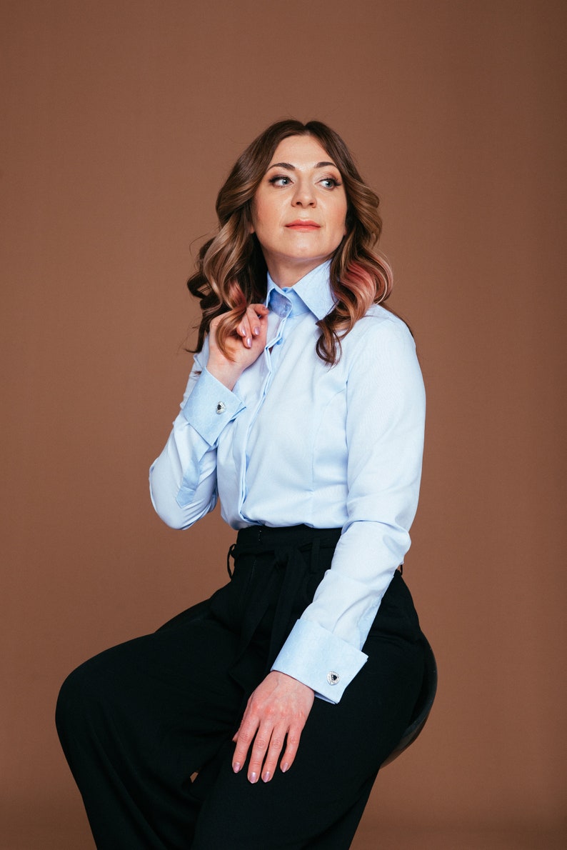 May include: A light blue button-down shirt with a contrasting white collar and cuffs. The shirt is paired with black wide-leg pants. The cuffs feature silver cufflinks. The model is seated on a stool against a brown backdrop.