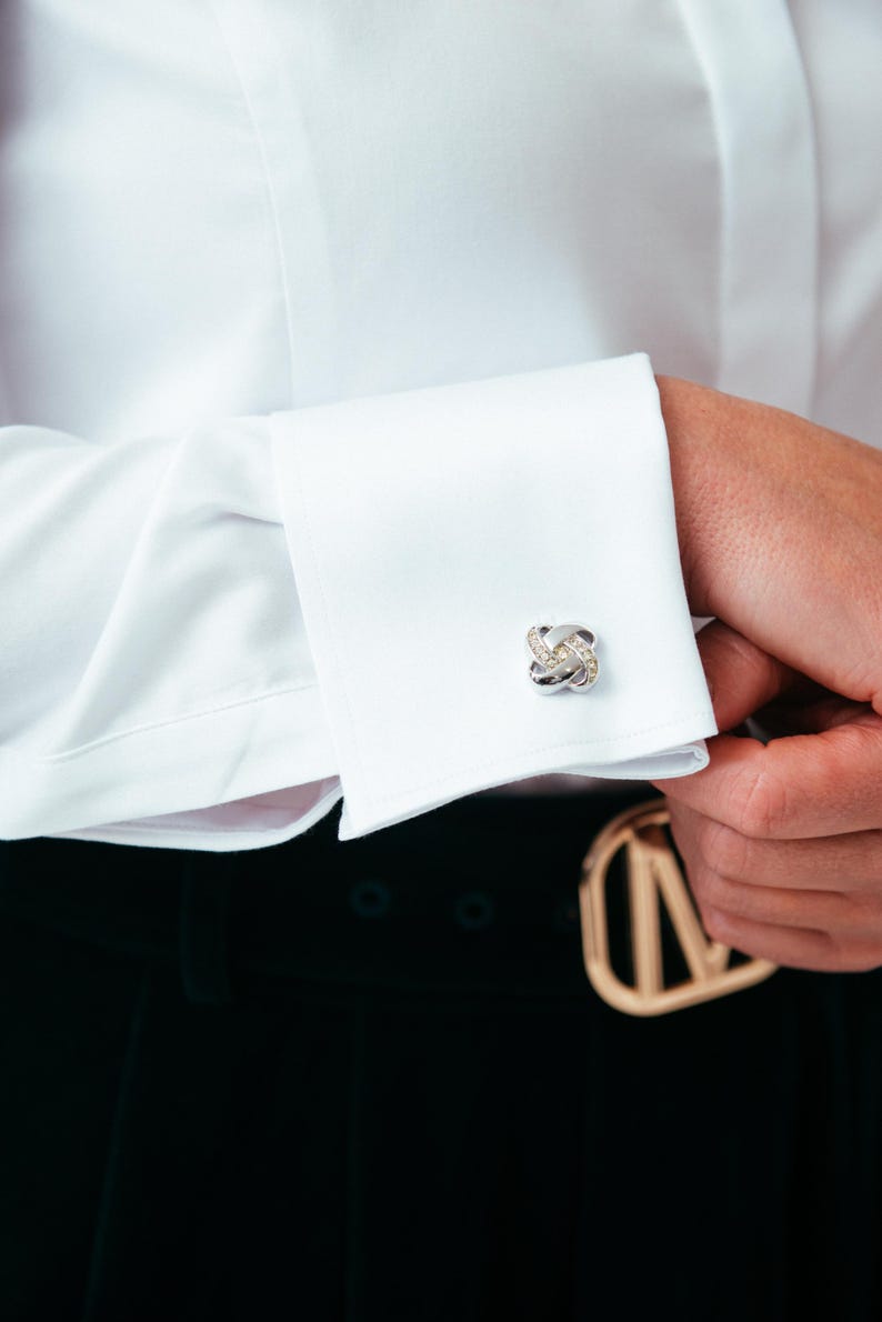 K&ouml;nnte beinhalten: Ein wei&szlig;es Langarmhemd mit Umschlagmanschette, die mit einem polierten, silberfarbenen Manschettenknopf in Knotenform verschlossen ist. Der Manschettenknopf hat ein zentrales, gedrehtes Design mit kleinen, klaren Steinen besetzt.