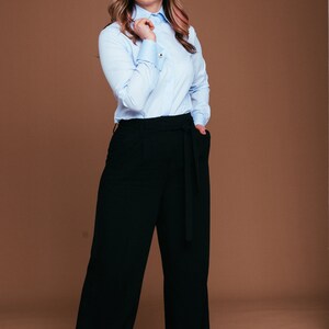 May include: A light blue button-down shirt with gold cuff links and black wide-leg pants with a matching belt. The outfit is a classic business casual style. The model is standing in front of a brown background.