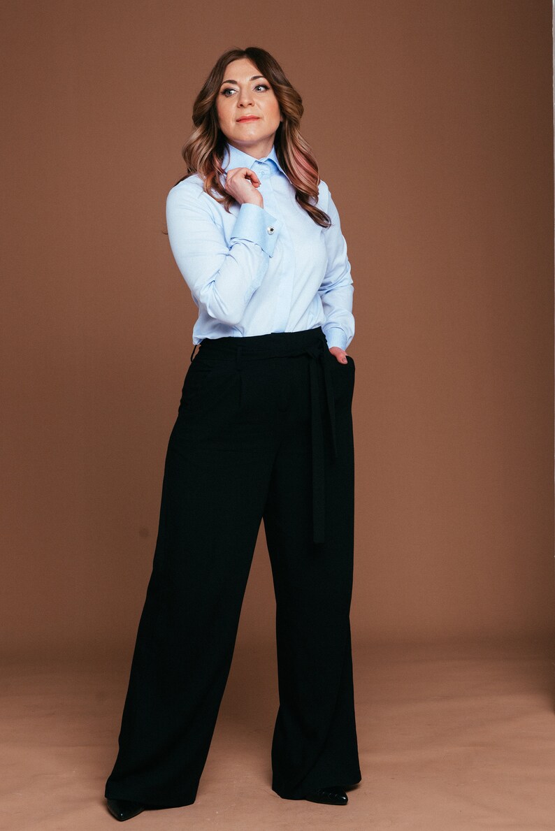 May include: A light blue button-down shirt with gold cuff links and black wide-leg pants with a matching belt. The outfit is a classic business casual style. The model is standing in front of a brown background.