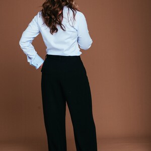 May include: A light blue button-down shirt with French cuffs and black wide-leg trousers. The outfit is a classic business casual style. The model is facing away from the camera, with long, wavy brown hair.