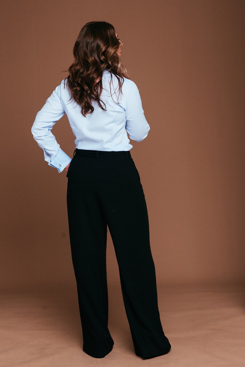 May include: A light blue button-down shirt with French cuffs and black wide-leg trousers. The outfit is a classic business casual style. The model is facing away from the camera, with long, wavy brown hair.