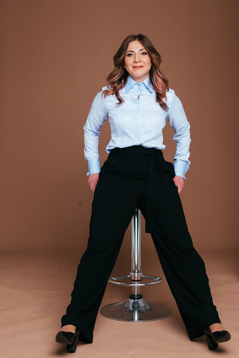 May include: A woman wearing a light blue button-down shirt with a classic collar and black wide-leg pants. The pants have a tie-waist detail. She is sitting on a chrome stool, with black heels. The background is a solid brown.