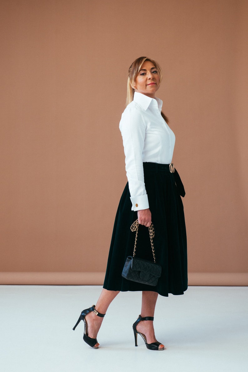 May include: A woman wearing a white button-down shirt with gold cufflinks, a black skirt with a gold belt, and black heels. She is carrying a black quilted handbag with a gold chain strap.