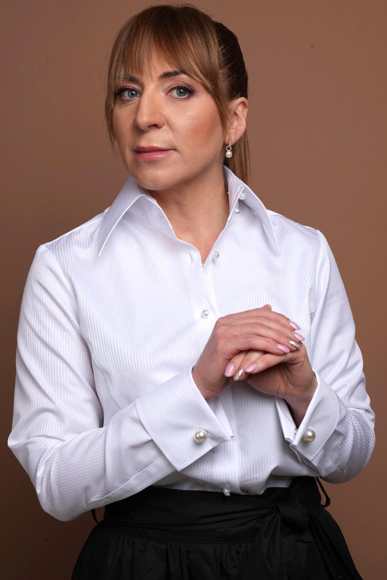 The white high collar stands tall and structured, giving the blouse a statement look. The fine cotton fabric is striped with subtle white-on-white detailing. Perfect for the confident woman looking for a versatile piece that combines comfort, prestige, and sophistication. Keywords: high collar blouse, white blouse, white cotton blouse.