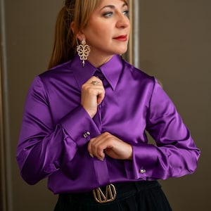 May include: A vibrant purple satin shirt with a classic collar and buttoned cuffs. The long-sleeved blouse is paired with a black skirt and a gold-toned belt. The image showcases a sophisticated, elegant style.