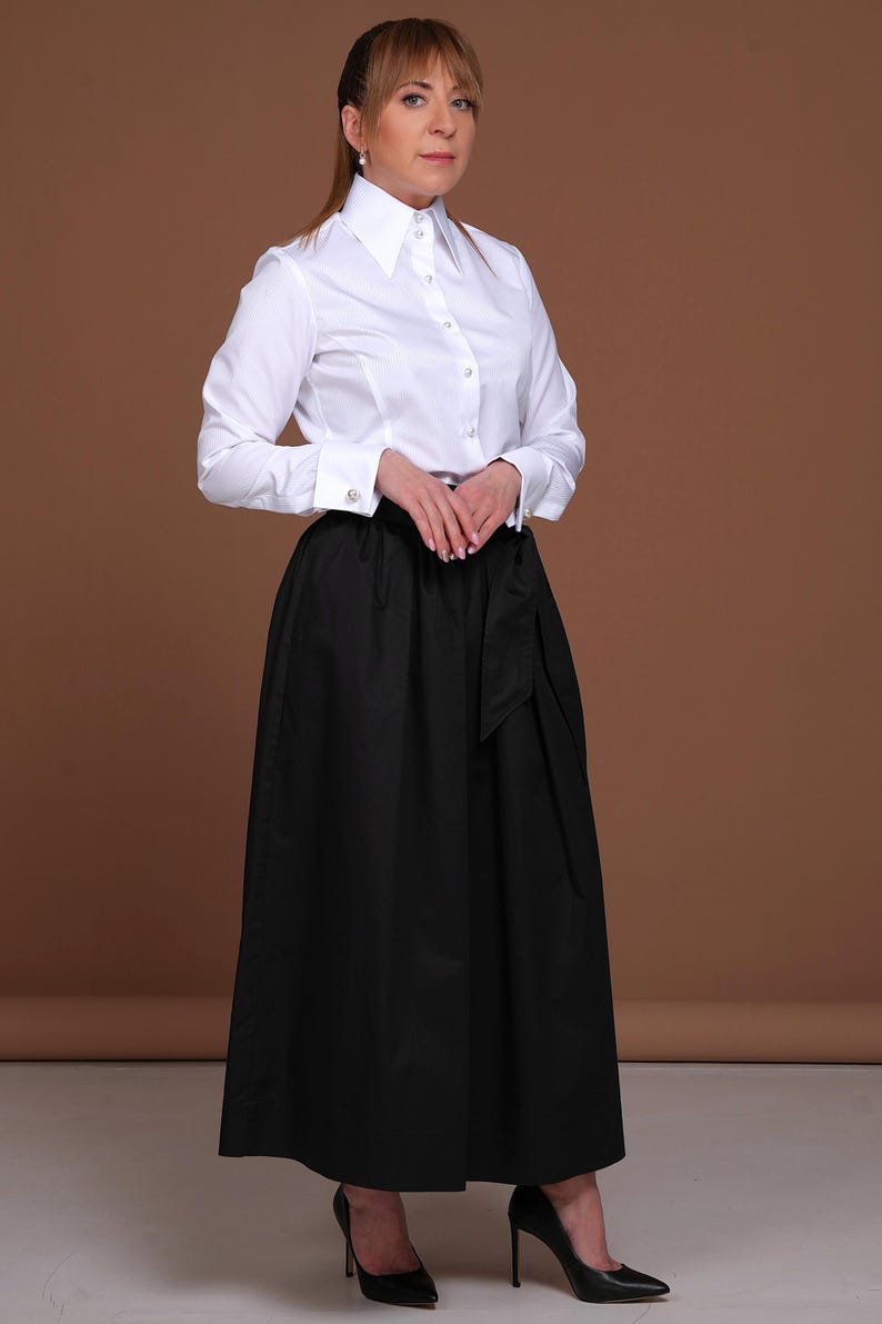 May include: A crisp white long-sleeved button-down shirt with a structured collar and cuffs, paired with a high-waisted black maxi skirt featuring a wide waistband and a tie-front detail. The ensemble is completed with black pointed-toe high heels.