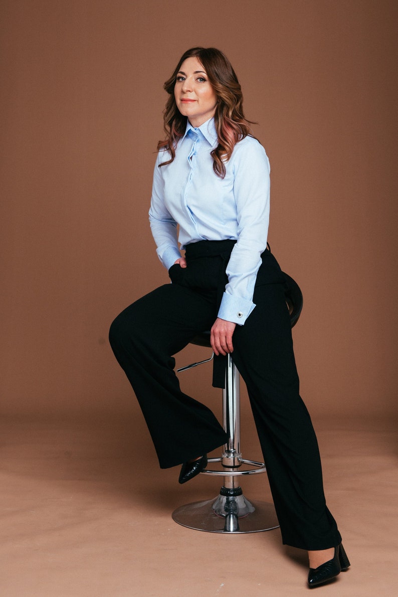May include: A woman wearing a light blue button-down shirt and black wide-leg pants. The shirt has long sleeves and a classic collar. She is seated on a chrome stool, with one hand in her pocket. Black pointed-toe heels complete the outfit.