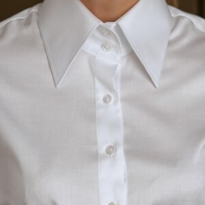 May include: Close-up of a crisp white button-down shirt. The shirt features a classic collar and a series of small, round buttons down the front. The fabric appears to be a smooth, woven material, suggesting a formal or professional style.