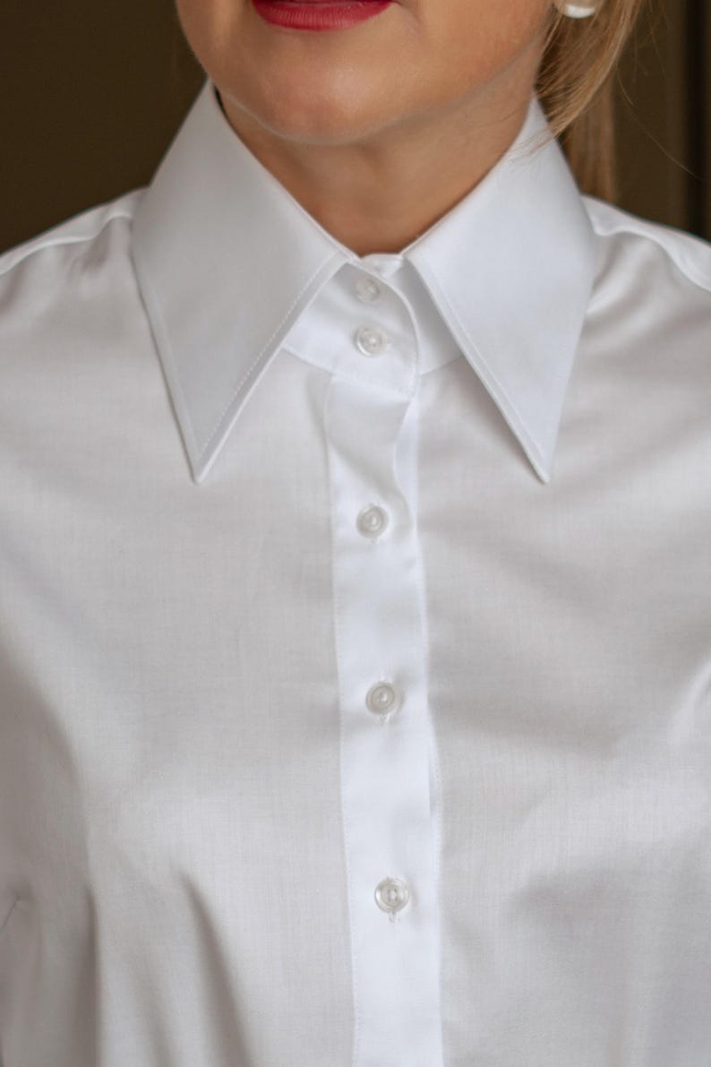 May include: Close-up of a crisp white button-down shirt. The shirt features a classic collar and a series of small, round buttons down the front. The fabric appears to be a smooth, woven material, suggesting a formal or professional style.
