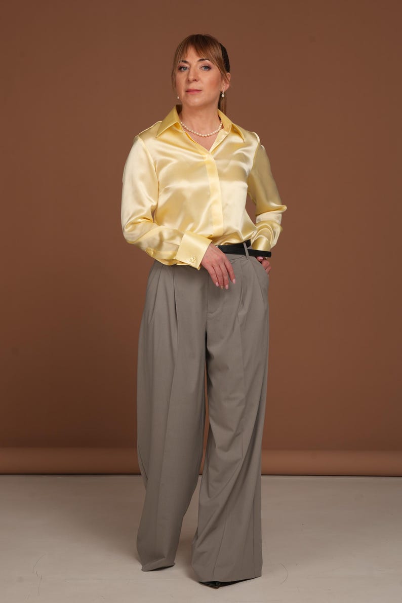 Full-body front view of a woman wearing a luxurious butter yellow silk blouse with French cuffs and a classic collar, styled with elegant wide-leg grey trousers. The smooth, high-quality silk fabric reflects light softly, emphasizing the premium craftsmanship and timeless silhouette of this elegant blouse