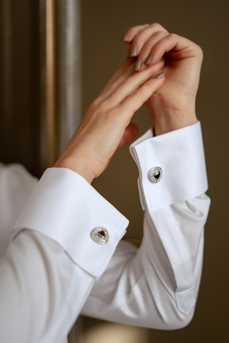 May include: Close-up of a white dress shirt with French cuffs. The cuffs are fastened with silver cufflinks featuring a black center stone surrounded by clear crystals. The image highlights the details of the formal wear.