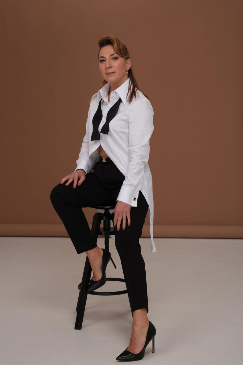 May include: A white button-down shirt with a black bow tie, paired with black trousers and heels. The outfit is completed with a black belt and gold accents. The model is seated on a black stool.