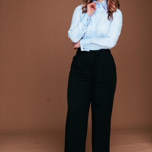 May include: A light blue button-down shirt with French cuffs and black wide-leg pants. The outfit is a classic business casual style. The pants have a matching belt. The background is a solid brown color.
