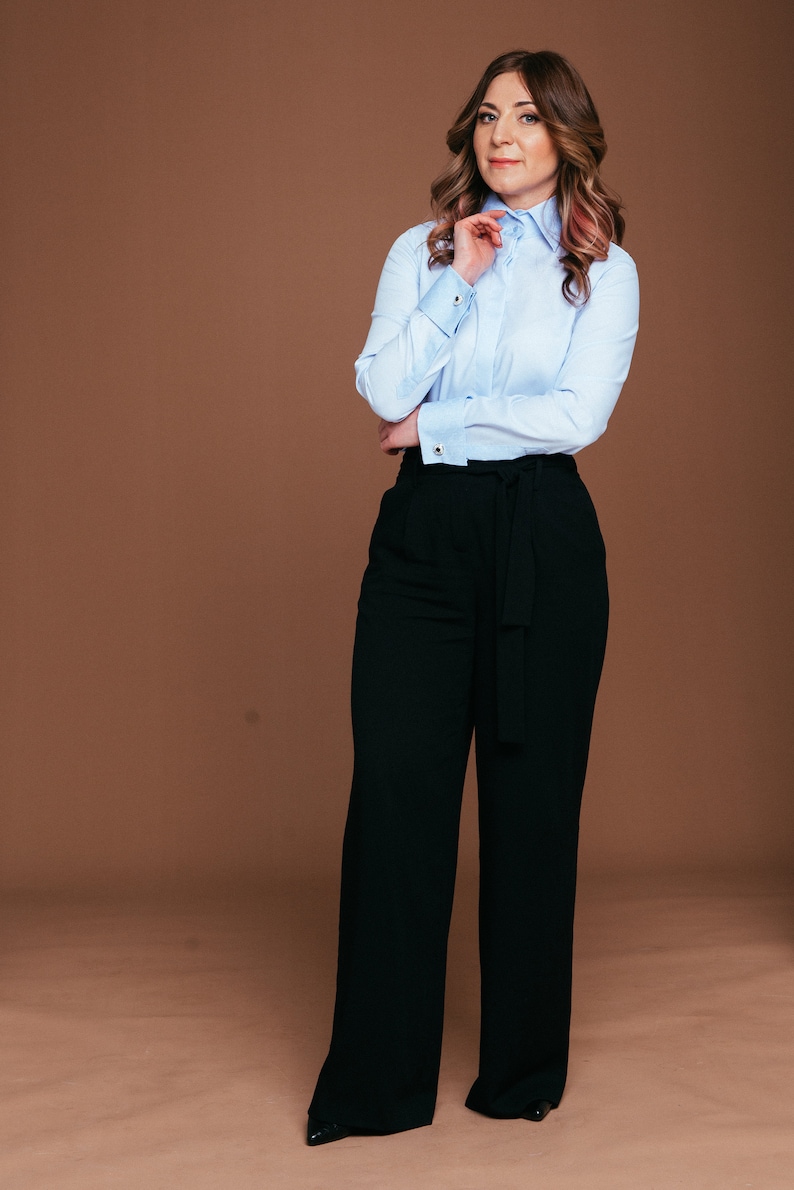 May include: A light blue button-down shirt with French cuffs and black wide-leg pants. The outfit is a classic business casual style. The pants have a matching belt. The background is a solid brown color.