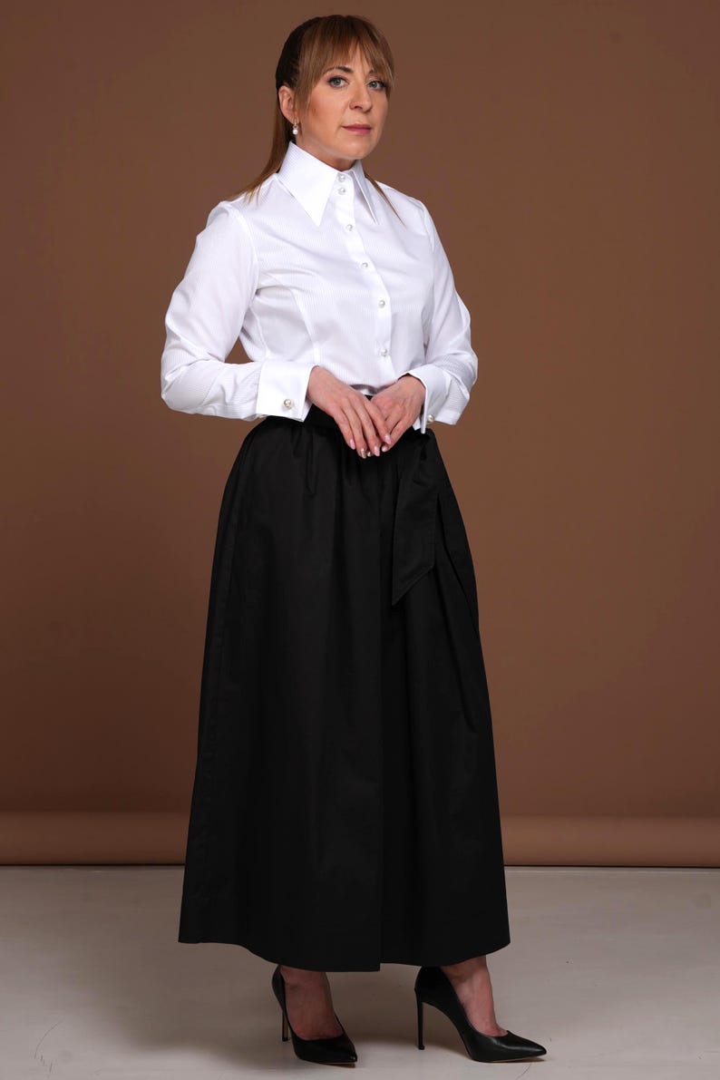 Elegant woman wearing a luxury white cotton blouse with fine vertical stripes, styled with a high collar and French cuffs for cufflinks. The fitted design emphasizes her silhouette, and the shirt is tucked into a voluminous black skirt for a refined, business-casual or formal look. The pearl buttons add timeless charm to this exclusive DaDonna Blouses model. Perfect for Google indexing: white blouse, white shirt women, French cuff blouse.