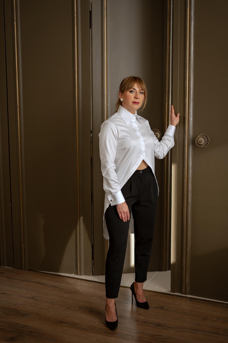 May include: A white, long-sleeve shirt with a unique asymmetrical hemline, paired with black trousers and black high heels. The shirt features a classic collar and button-down front. The outfit is a stylish ensemble.