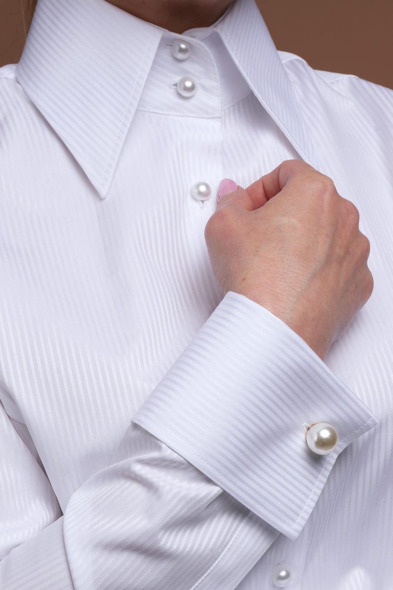 Close-up portrait showing the elegant white blouse with high standing collar and luxury pearl buttons. The crisp cotton fabric has a delicate striped texture, making it ideal for women seeking a sophisticated and timeless business or evening look. Styled with drop pearl earrings for an extra touch of class. Keywords: white top, white formal blouse, French cuff blouse